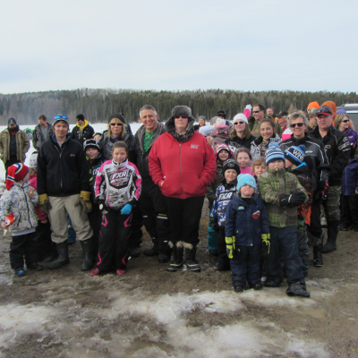 This photo depicts a large group of men, women and children who participated in the Family Ice Fishing Derby held in Hornepayne. They gathered together for a group photo on the ice with the lake behind them.