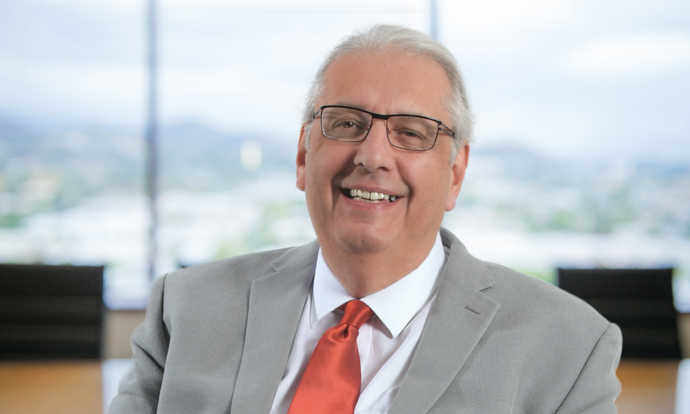 Picture shows Bob Watts smiling, with an office backdrop that includes large windows with a city view behind him.