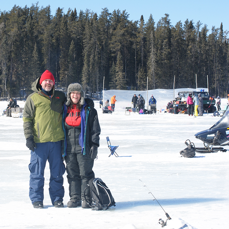 Two people stand on the snow, with winter activities in the background