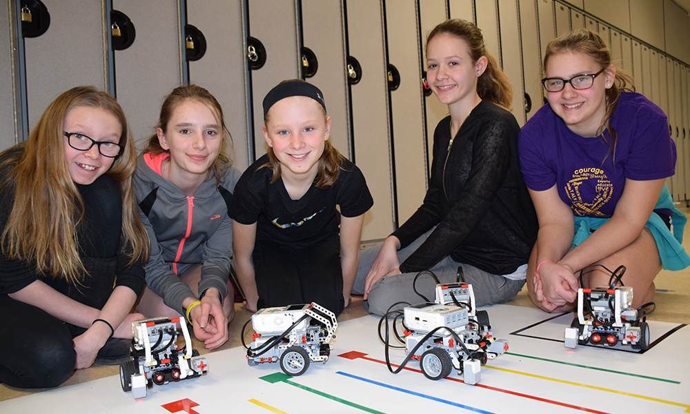 The photo is of five Grades 7 and 8 girls, side by side, on the floor with robots constructed out of LEGO kits by themselves and fellow classmates.