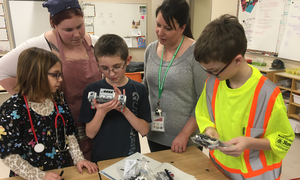 Trois écoliers essaient les ensembles de robotique sous la sourveillance de deux adultes 