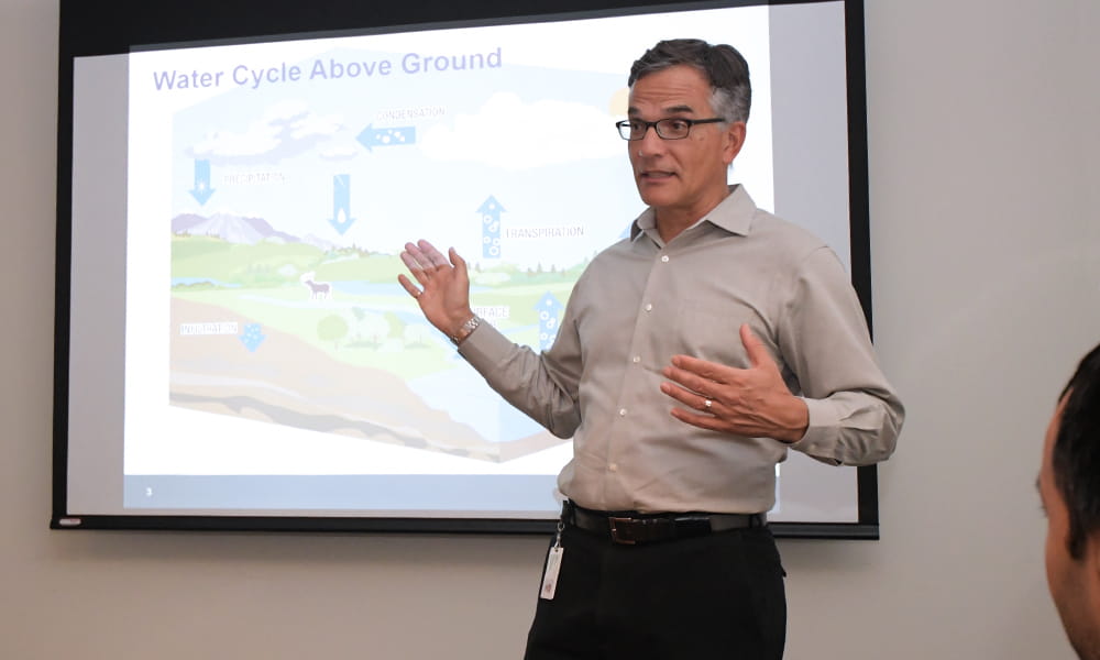 Photo shows a man giving a presentation in front of a projection screen.