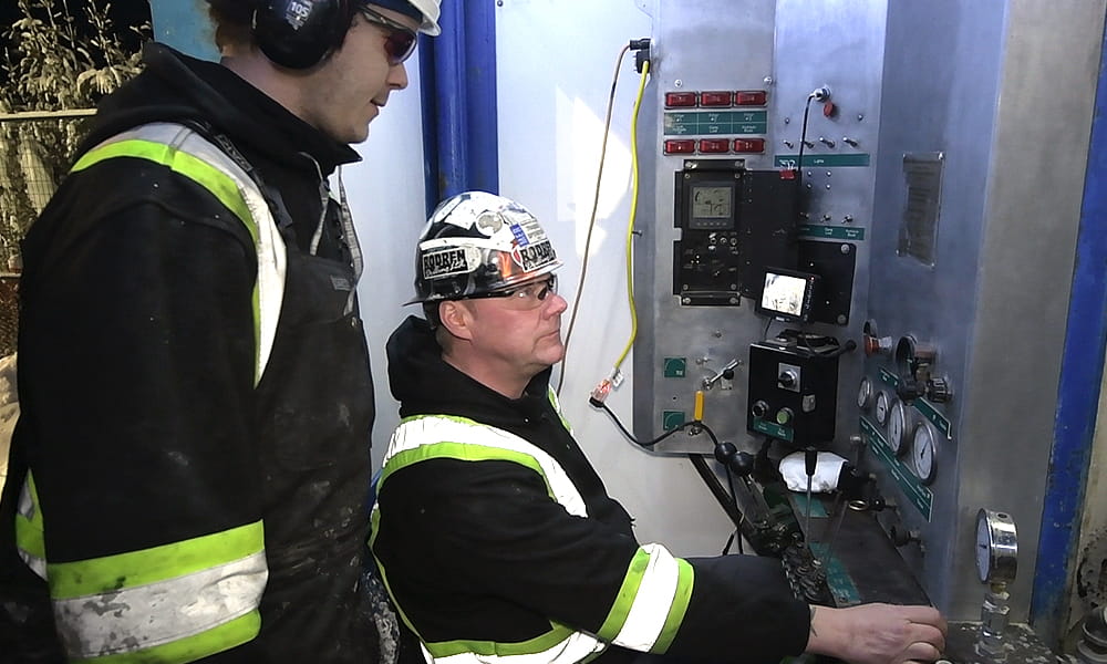 Un foreur travaille assis accompagné par son collègue debout, derrière lui