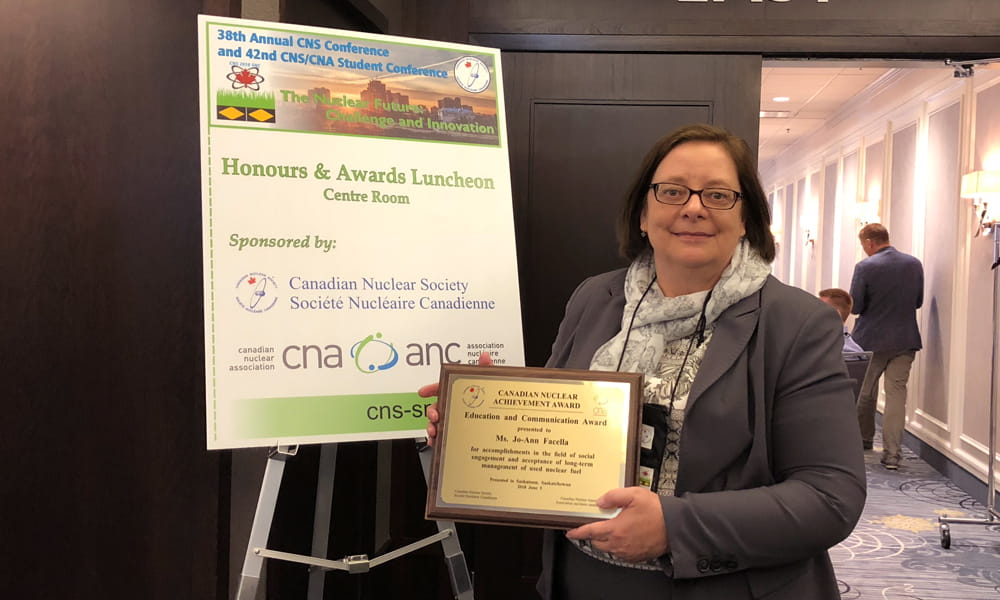 Image shows a woman, Jo-Ann Facella, standing with her award, which is a plaque with an inscription. 