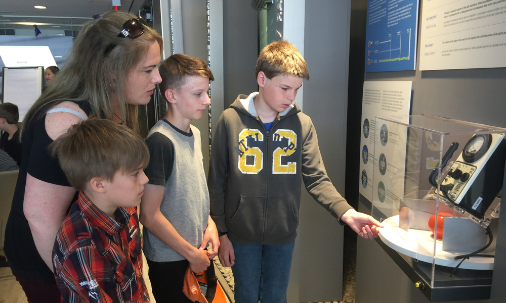 Image shows three young students with their teacher looking at an unspecified display item. 