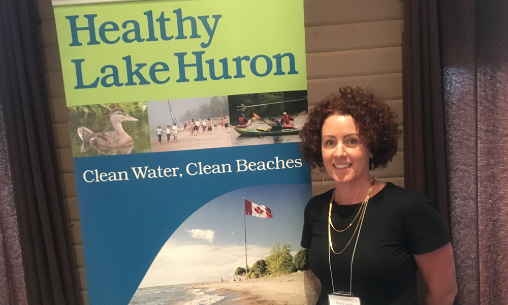 A woman standing in front of a “Healthy Lake Huron” poster.