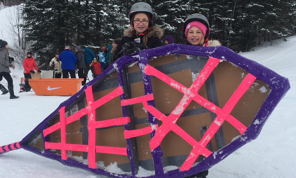 Photo depicts two young residents standing on snow with their colourful snowshoe-themed cardboard box racer.