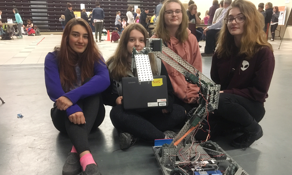 Image shows four girls sitting behind their robotic creation. 