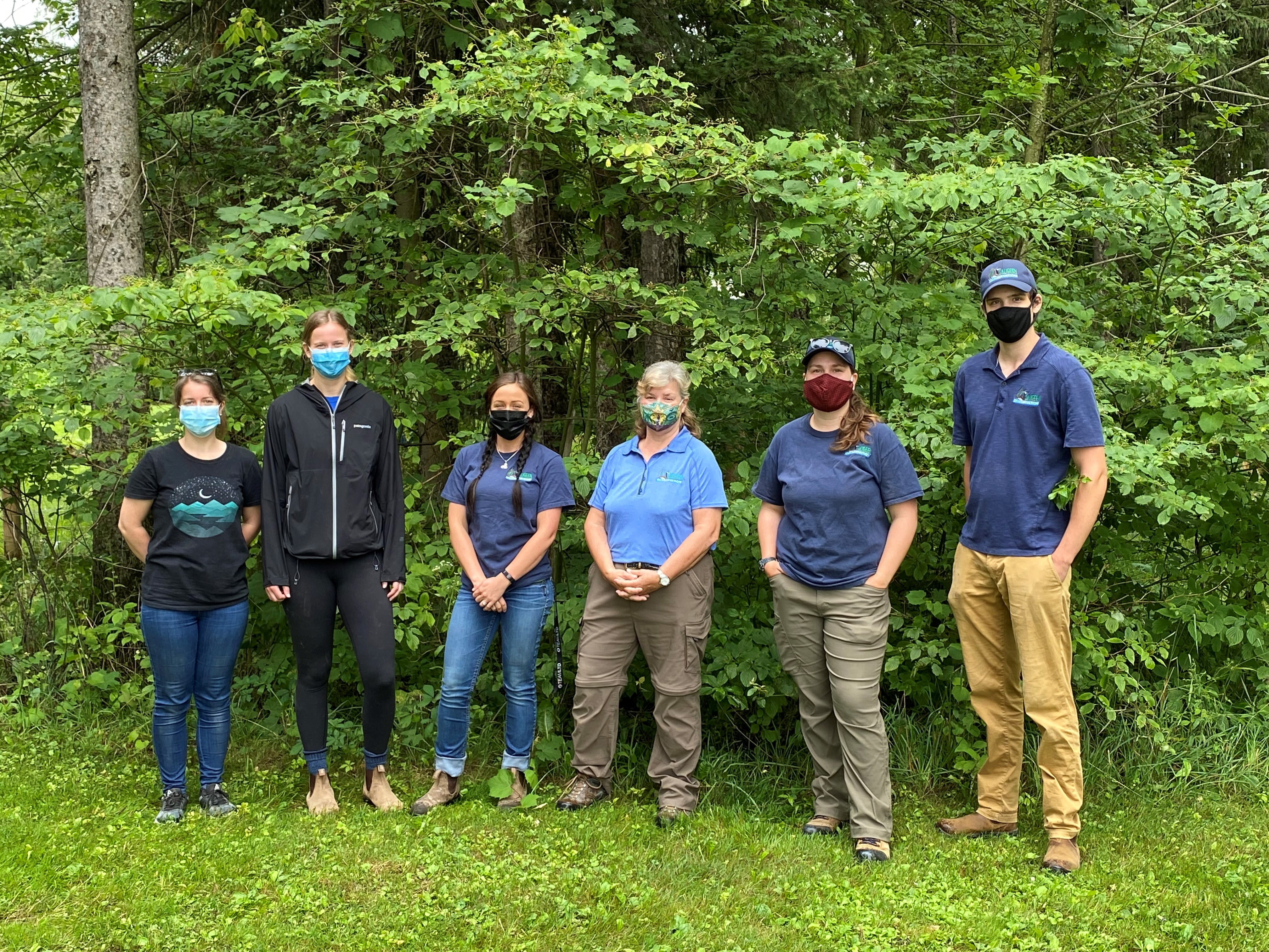 Six individuals (five women and one man) standing side-by-side on lawn in front of some bushes and trees. They are all wearing face masks.